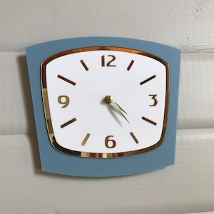 May include: A blue and gold wall clock with a white face and black hour and minute hands. The clock has Roman numeral markings for the hours and Arabic numerals for the 3, 6, 9, and 12.