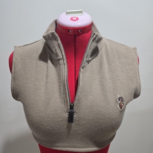May include: A tan-colored, sleeveless sweater with a high collar and a zipper down the front. The sweater has a small emblem on the chest. The garment is displayed on a red dress form.