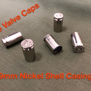 Puede incluir: Cinco tapones de válvula de neumático plateados y brillantes hechos de casquillos de níquel de 9 mm. Los tapones están dispuestos sobre una superficie de tela verde. El texto de la imagen dice "Tire Valve Caps" y "9mm Nickel Shell Casing."