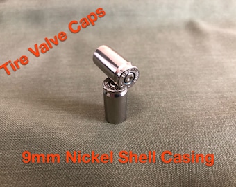 May include: Two silver-colored tire valve caps made from 9mm nickel shell casings. The caps are cylindrical with a detailed top and are displayed on a green fabric surface. Text on the image reads "Tire Valve Caps" and "9mm Nickel Shell Casing."