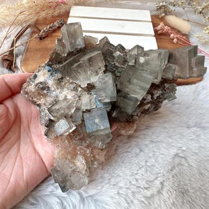May include: A hand holds a cluster of grey and clear cubic crystals, possibly fluorite. The crystals have a geometric shape and are clustered together. The background includes a wooden board and decorative elements.