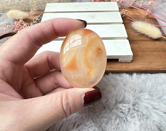 May include: A smooth, polished carnelian stone with a light orange and white banded pattern. The stone is oval shaped and has a glossy finish.