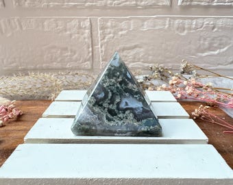May include: A green and white agate pyramid crystal. The pyramid is sitting on a white surface.