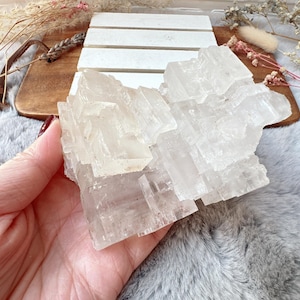 May include: A large, clear selenite crystal cluster. The translucent mineral has a layered, geometric structure. The selenite is held in a hand, with a wooden board and decorative elements in the background. The crystal is a natural, earthy item.