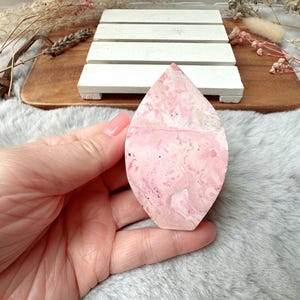 May include: A pink, teardrop-shaped stone with a marbled pattern of lighter pink and white. The stone is held in a hand, with a wooden tray and white slatted platform in the background. Dried flowers and greenery are also visible.