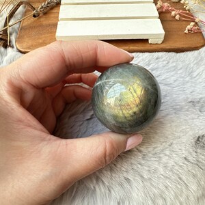 May include: A polished labradorite sphere, held in a hand. The sphere exhibits a play of colours, including blues, greens, and golds. The background includes a wooden tray, a white platform, and decorative elements.