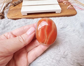May include: A polished, oval-shaped gemstone with bands of orange and white. The stone is held in a hand, showcasing its smooth, reflective surface. The gemstone is likely a decorative item or a piece of jewelry.