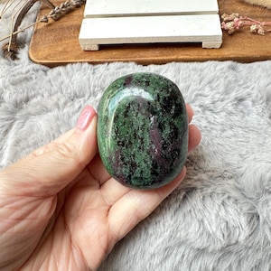 May include: A polished, oval gemstone displaying a mix of green and dark red colours. The stone has a smooth, glossy surface and is held in a hand. The gemstone is a natural, decorative item.