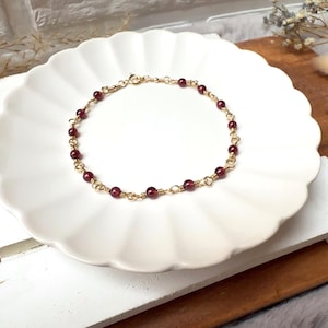 May include: A gold-colored bracelet with small, round, dark red beads. The bracelet is resting on a white, scalloped-edge plate. The background includes dried flowers and a wooden surface.