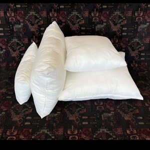 May include: Four white pillow inserts of different sizes. The pillows are arranged in a stack on a dark patterned surface.