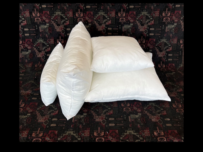 May include: Four white pillow inserts of different sizes. The pillows are arranged in a stack on a dark patterned surface.