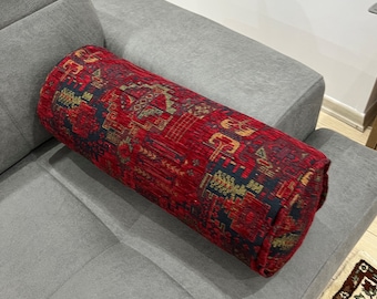 May include: A cylindrical decorative pillow with a red, navy, and gold patterned design. The pillow is resting on a gray sofa. The pattern appears to be a traditional rug design.