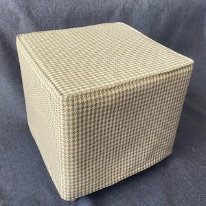 May include: A square ottoman with a beige and brown houndstooth pattern fabric cover.