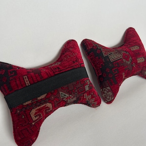 May include: Two red and black patterned neck pillows with a black elastic band. The pillows are shaped like a V and are made of a soft, plush fabric.