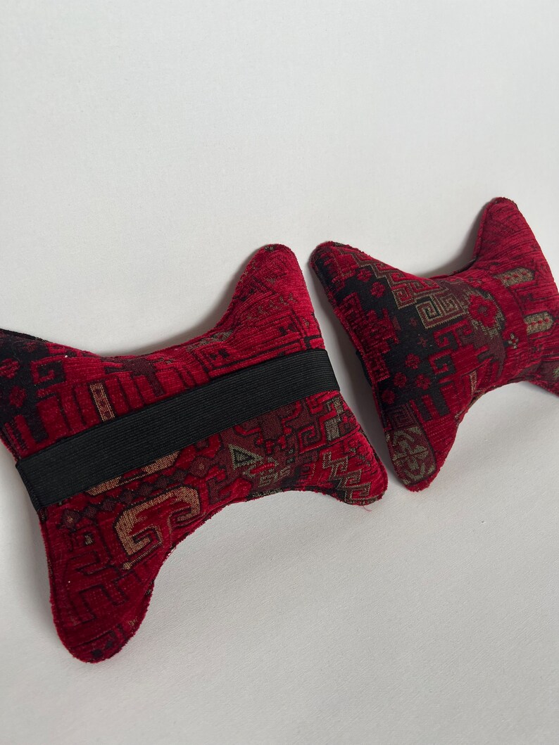 May include: Two red and black patterned neck pillows with a black elastic band. The pillows are shaped like a V and are made of a soft, plush fabric.