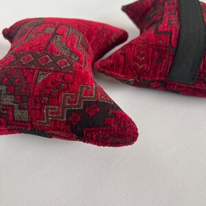 May include: Two red and black patterned throw pillows with black elastic bands. The pillows have a geometric design.