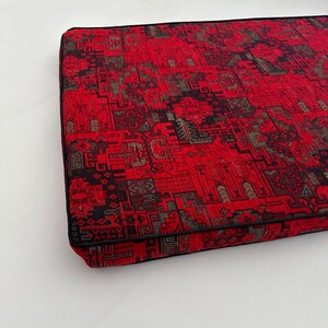 Puede incluir: Coj&iacute;n rojo y negro con estampado y borde negro. El coj&iacute;n est&aacute; hecho de una tela suave y texturizada.