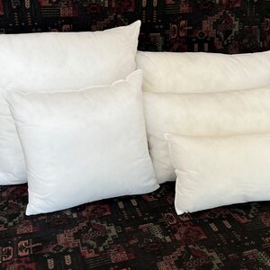 May include: Five white pillow inserts of varying sizes stacked on top of each other. The pillows are arranged in a pyramid shape with the largest pillow at the bottom and the smallest pillow at the top.