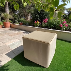 May include: A beige square ottoman with a cushioned top sits on artificial green grass. The ottoman has rounded edges and a light brown trim. The background features a patio, plants, and flowers, suggesting an outdoor setting.