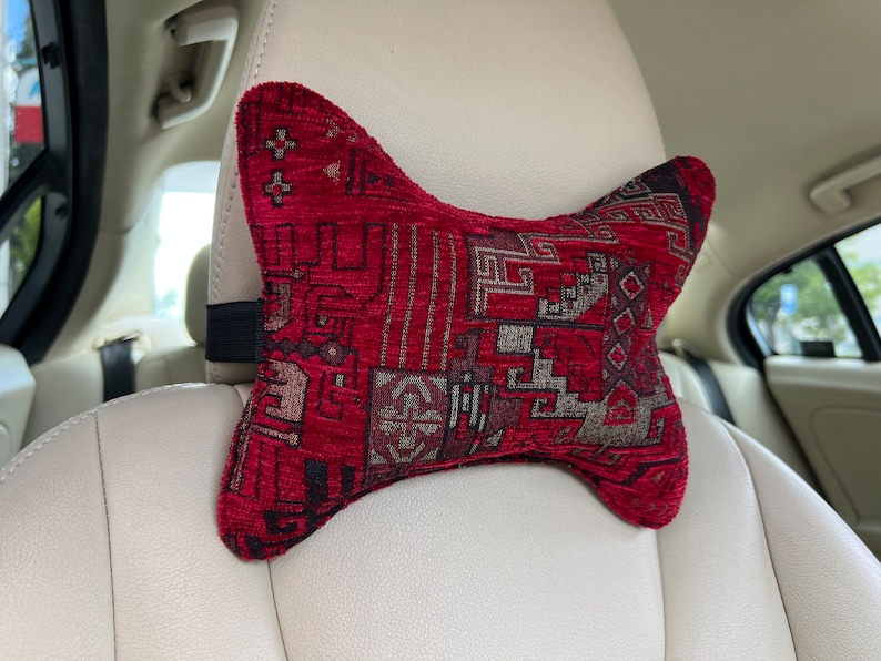 May include: A red and gray patterned car headrest pillow with a strap to attach it to the headrest.