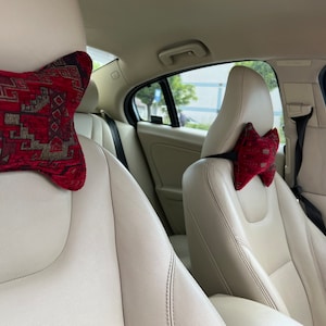May include: Two car seats with beige leather upholstery and red patterned headrest pillows. The pillows are shaped like a bow tie.