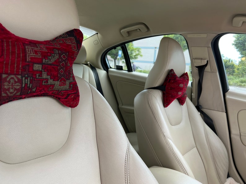 May include: Two car seats with beige leather upholstery and red patterned headrest pillows. The pillows are shaped like a bow tie.