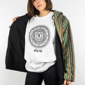 Op de afbeelding: Een wit t-shirt met een zwart mandala-ontwerp en tekst, gedragen onder een zwarte jas met ritssluiting met een kleurrijk gestreept patroon op de mouwen. De persoon draagt een zwarte broek.