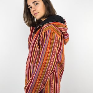 Può includere: Una donna indossa una giacca con cappuccio a righe colorate con un motivo geometrico. La giacca ha una cerniera e un cappuccio con coulisse.