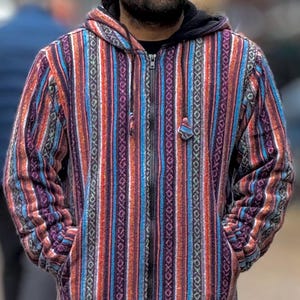 Könnte beinhalten: Ein Mann trägt eine gestreifte, mehrfarbige Kapuzenjacke mit Reißverschluss. Die Jacke hat eine dunkelblaue, rote, orange und violette Farbgebung. Er trägt auch eine rote Baseballkappe und eine blaue Jeans.