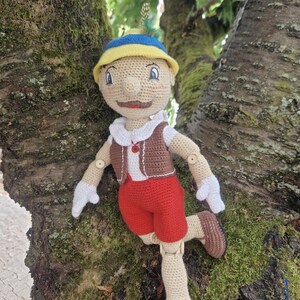 May include: A crocheted doll with a yellow hat, brown vest, and red pants. The doll is standing on a tree trunk with green moss.