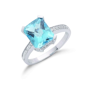 May include: A silver ring featuring a large, rectangular, light blue gemstone. The band is adorned with small, clear stones. The ring is set against a white background, highlighting its elegant design.