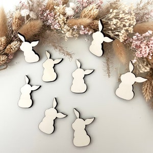 Peut inclure: Huit formes de lapin en bois blanc avec des contours noirs, disposées sur une surface blanche avec des fleurs séchées en arrière-plan.