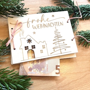 May include: A wooden Christmas card with the words "Frohe Weihnachten" engraved on it, along with a house and Christmas tree design. A pink string is used to tie the card together. A five Euro bill is visible.