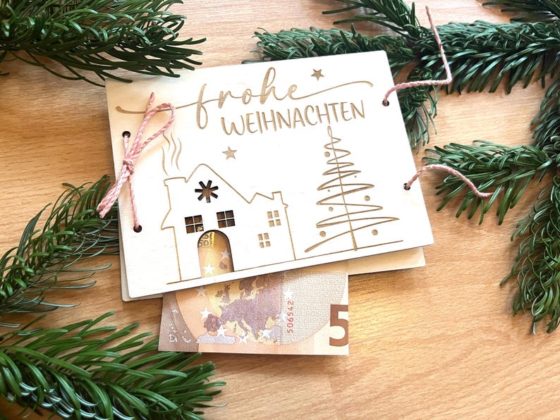May include: A wooden Christmas card with the words "Frohe Weihnachten" engraved on it, along with a house and Christmas tree design. A pink string is used to tie the card together. A five Euro bill is visible.