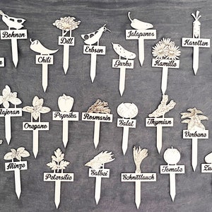 Könnte beinhalten: Eine Sammlung weißer Gartenstecker aus Holz mit verschiedenen Gemüse- und Kräuterillustrationen und -namen. Die Stecker tragen Namen wie "Basilikum", "Chili", "Oregano" und "Tomate". Die Stecker sind auf einer grauen Oberfläche angeordnet.