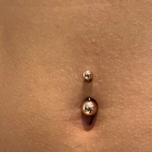 May include: A gold-colored belly button ring with two spherical ends. The larger ball is at the bottom, and the smaller ball is at the top. The skin around the piercing is visible.