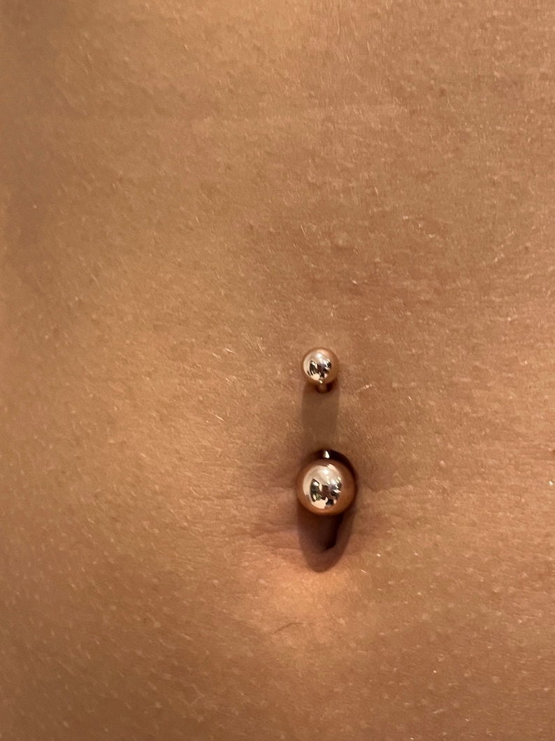 May include: A gold-colored belly button ring with two spherical ends. The larger ball is at the bottom, and the smaller ball is at the top. The skin around the piercing is visible.