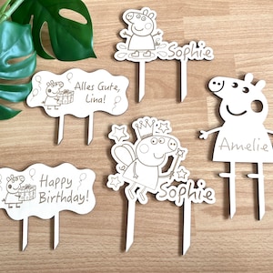 Könnte beinhalten: Vier weiße Holz-Kuchentopper mit einem Comic-Schweinecharakter. Die Topper zeigen ein Schwein mit einer Krone, ein Schwein, das ein Geschenk hält, ein Schwein mit Ballons und ein Schwein, das mit dem Text "Amelie" steht.