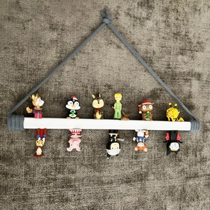 Op de afbeelding: Een wit houten plank met een grijs touw dat aan het plafond hangt. De plank is versierd met verschillende kleine beeldjes, waaronder een pingu&iuml;n, een eekhoorn, een prins, een sneeuwpop, een bij, een vleermuis en een cartoonfiguur.