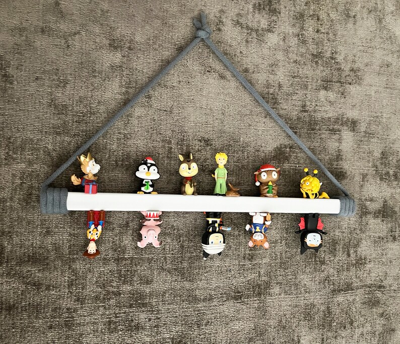 Op de afbeelding: Een wit houten plank met een grijs touw dat aan het plafond hangt. De plank is versierd met verschillende kleine beeldjes, waaronder een pingu&iuml;n, een eekhoorn, een prins, een sneeuwpop, een bij, een vleermuis en een cartoonfiguur.