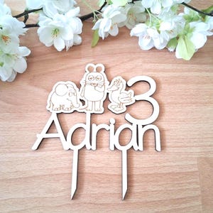 Könnte beinhalten: Kuchenaufsatz aus Holz mit dem Namen "Adrian" und der Zahl "3". Der Aufsatz zeigt Cartoon-Tierdesigns eines Elefanten, eines Kaninchens und einer Ente. Der Aufsatz ist aus hellem Holz gefertigt.