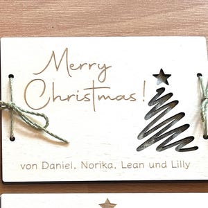 May include: A wooden Christmas card with the words "Merry Christmas!" in elegant script. A star and a stylized Christmas tree are cut out of the wood. The card is tied with green twine.