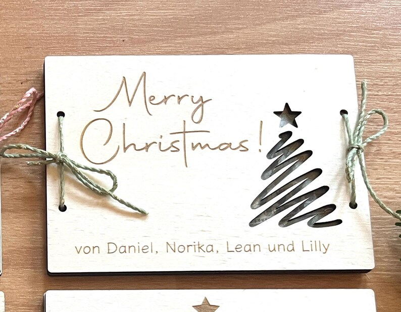 May include: A wooden Christmas card with the words "Merry Christmas!" in elegant script. A star and a stylized Christmas tree are cut out of the wood. The card is tied with green twine.