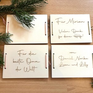 May include: Four wooden gift tags with handwritten text in brown ink. The tags are rectangular with string and pink thread accents. Text includes phrases like "For Mirian" and "For the best Grandma in the world". Green pine branches are in the background.