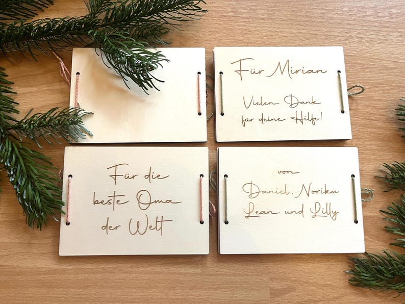 May include: Four wooden gift tags with handwritten text in brown ink. The tags are rectangular with string and pink thread accents. Text includes phrases like "For Mirian" and "For the best Grandma in the world". Green pine branches are in the background.