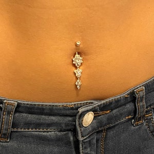 May include: A gold belly button ring with three dangling charms, each featuring a cluster of clear rhinestones.