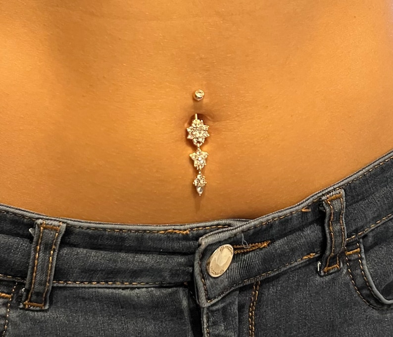 May include: A gold belly button ring with three dangling charms, each featuring a cluster of clear rhinestones.