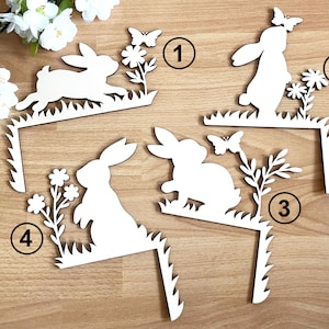 Könnte beinhalten: Vier weiße Holz-Hasensilhouetten mit Blumen und Schmetterlingen. Jeder Hase ist an einem Holzständer mit grasartigem Design befestigt. Die Ständer sind von 1 bis 4 nummeriert.