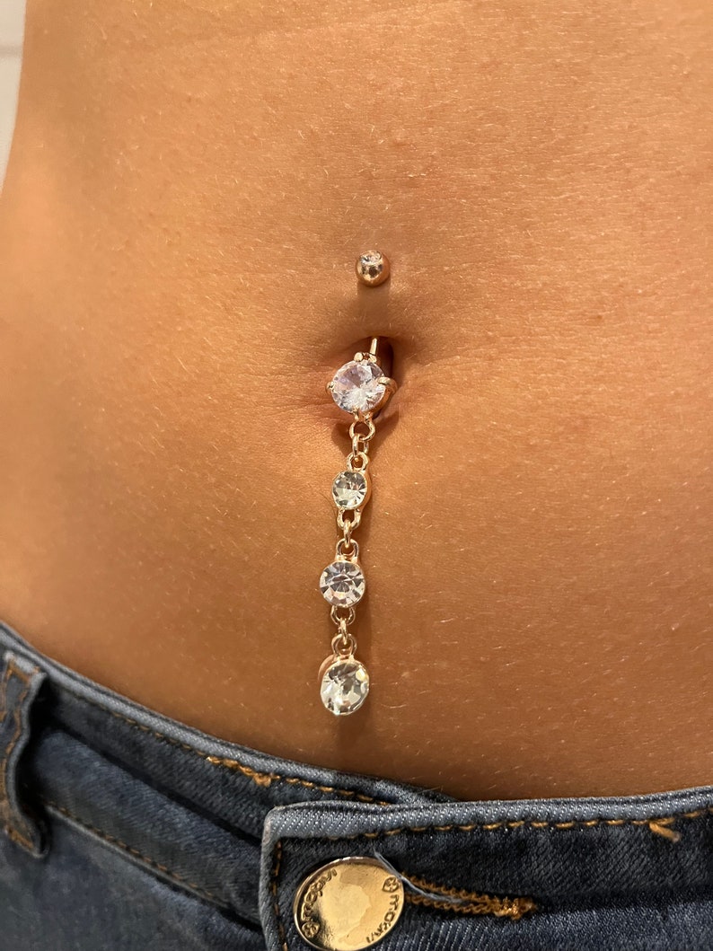 K&ouml;nnte beinhalten: Ein goldener Bauchnabelpiercing mit einer baumelnden Kette und klaren Kristall-Akzenten.