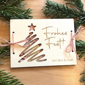 May include: A wooden Christmas card with a cut-out Christmas tree design and a star at the top. The card is tied with pink twine and has the words "Frohes Fest!" and "von Ben & Axel" engraved on it. Green pine branches surround the card.